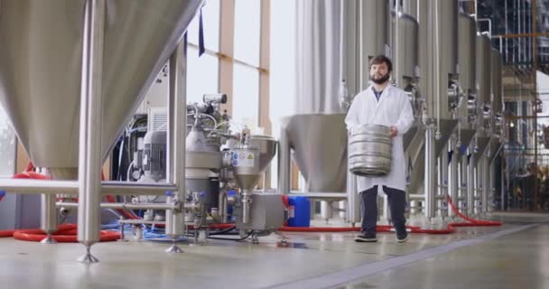 Een werkende mannelijke brouwer in laboratoriumjas draagt een vat vol bier voor zich als hij langs biertanks loopt. Langzame beweging — Stockvideo
