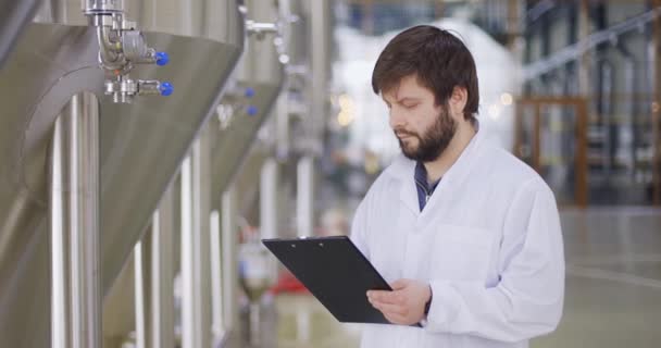 Hombre con bata de laboratorio blanca mirando el medidor del equipo e ingresando datos en la tableta. Trabajador de cervecería macho parado en filas inoxidables de cubas de cerveza — Vídeos de Stock