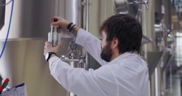 Brewery worker in work process. Man dressed in lab coat at brewery factory near beer tanks — Stock Video