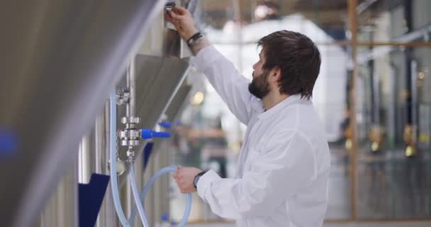 Brouwerij werknemer in het arbeidsproces. Man gekleed in labjas in brouwerij fabriek in de buurt van biertanks — Stockvideo