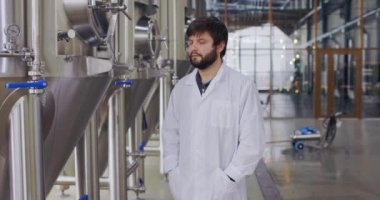 Fabrikadaki kameraya poz veren laboratuvar önlüklü genç teknoloji uzmanının portresi. Bira fabrikasının arka planında bira üreticisi.
