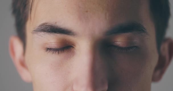 Крупный план спокойного красивого молодого человека открывает глаза и смотрит в камеру на сером фоне в замедленной съемке — стоковое видео