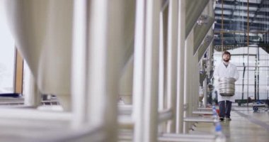 Laboratuvar önlüklü bir işçi bira tanklarının yanından geçerken önünde bira dolu bir fıçı taşıyor. Yavaş çekim