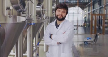Laboratuvar önlüklü genç teknoloji uzmanının portresi bira fabrikasında kameraya poz veriyor. Bira fabrikasının arka planında bira üreticisi.