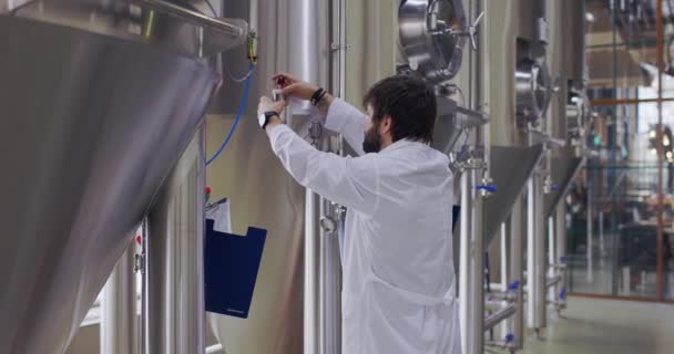 Trabajador cervecero en proceso de trabajo. Hombre vestido con bata de laboratorio en la fábrica de cerveza cerca de tanques de cerveza — Vídeos de Stock