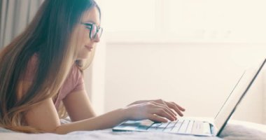 Yatak odasında dizüstü bilgisayarında daktilo yazan gözlüklü bir kadın. Parlak güneşli ışık