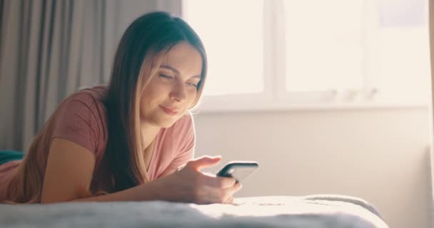 幸せな若い女性がベッドの上に横たわって、午前中に自宅で電話を使用しています。明るい陽射し — ストック動画