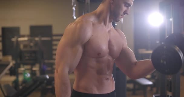 Sportovní mladík bez trička, který cvičí s činkou v tělocvičně. Pohlední muži pumpují biceps svaly. Zpomalený pohyb — Stock video