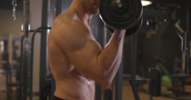 Spor salonunda halterle egzersiz yapan atletik tişörtsüz genç bir sporcu. Yakışıklı adamlar kaslarını şişiriyor. Yavaş çekim