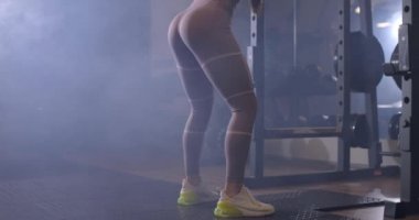 Spor giyimli güzel bir bayan karanlık bir spor salonunda halterle çömeliyor. Arka plan, yavaş çekim. Güç ve motivasyon kavramı