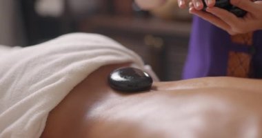Masör kaplıcadaki genç, çekici ve çekici bir kadının sırtına sıcak taşlar koyuyor. Çamaşır taşı terapisi. Spa ve sağlık konsepti. Yavaş çekim