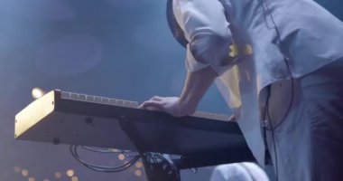 Genç bir adam sahnede sentezleyici çalıyor. Rock grubu konseri, neon ışıklar. Yavaş çekim