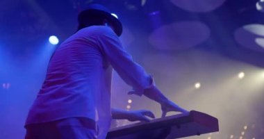 Genç bir adam sahnede sentezleyici çalıyor. Rock grubu konseri, neon ışıklar. Yavaş çekim