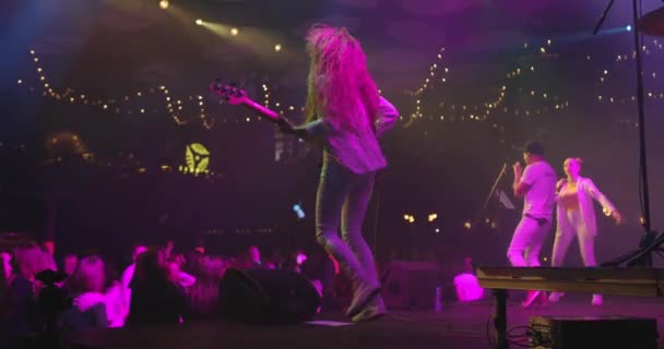 Rückansicht einer Gitarristin in weißer Kleidung mit Rockband bei einem Live-Konzert. Der Gitarrist hüpft mit der Bassgitarre und schüttelt seine Haare auf der Bühne. Zeitlupe — Stockvideo