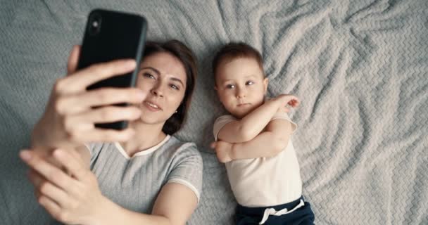 Anya és a kisfia szelfit vagy videót készítenek apának vagy rokonainak az ágyban. Technológia, új generáció, család, kapcsolat, szülőség. Felülnézet, lassított felvétel. — Stock videók