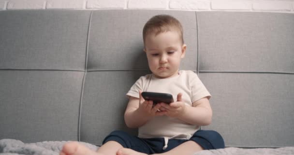 El niño se sienta en la cama sosteniendo un teléfono negro y mirándolo. El concepto de usar gadgets por los niños. Primer plano, cámara lenta — Vídeo de stock