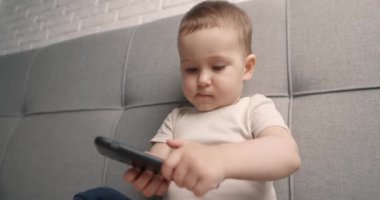 Küçük bebek yatakta oturuyor ve akıllı telefonuyla oynuyor. Siyah bir cep telefonu tutan bebekler. Yakın çekim, yavaş çekim