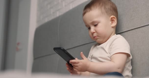 Kleine jongen zit op het bed naar de smartphone te kijken en te glimlachen. De baby 's met een zwarte gsm. Langzame beweging — Stockvideo
