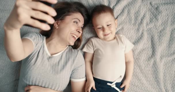 Gülen kadın ve bebek yatakta selfie ya da video çekiyor. Teknoloji kavramı, yeni nesil, aile, bağlantı, ebeveynlik. Üst görünüm, yavaş çekim. — Stok video