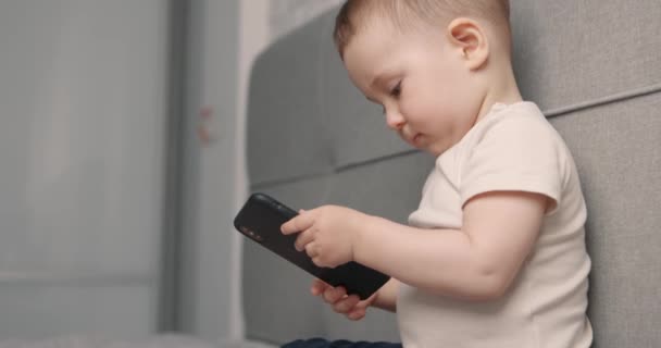 Rallentatore panning colpo di bel bambino tiene smartphone in mano e lo guarda seduto sul letto — Video Stock