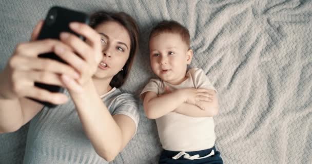 Мама и сын делают селфи с помощью мобильного телефона. Женщина и маленький мальчик лежат на кровати и смотрят в камеру смартфона. Видеозвонок. Вид сверху, замедленное движение — стоковое видео