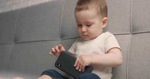 Повільний постріл красивого маленького хлопчика тримає смартфон в руках і дивиться на нього, сидячи на ліжку — стокове відео