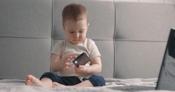Kleine jongen die op bed zit met een mobieltje in handen en aan het spelen. De laptop is vlakbij. Langzame beweging. — Stockvideo
