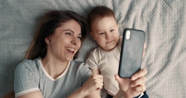 母親は携帯電話を使って赤ちゃんと一緒に自撮り。女性と子供はベッドの上に横になり、スマートフォンを使ってビデオ通話をします。スローモーション — ストック動画