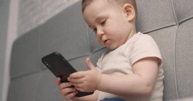 Güzel küçük oğlan akıllı telefonu elinde tutuyor ve yatakta otururken ona bakıyor. Bebek ekranda dokunuyor. Yan görünüm, yavaş çekim