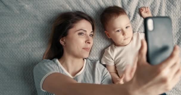 Usmívající se žena a chlapeček dělají selfie nebo video hovor v posteli a mávají rukama. Koncept technologie, nová generace, rodina, spojení, rodičovství. Pohled shora, zpomalený pohyb. — Stock video