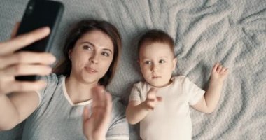 Anne, oğluyla cep telefonu kullanarak selfie çekiyor. Kadın ve çocuk yatağa uzanıp akıllı telefondan görüntülü arama yapıyorlar. Yavaş çekim