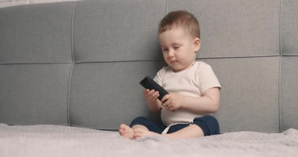 El niño se sienta en la cama y mantiene el control remoto desde el televisor. El niño enciende la televisión y sonríe. Movimiento lento — Vídeo de stock