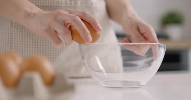 Wanita dengan celemek abu-abu mengambil telur dari keranjang dan memecahkannya menjadi mangkuk kaca. Memasak di dapur rumah. Gerakan lambat — Stok Video