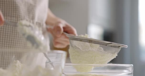 Жінка в сірому фартусі натирає сир ситом на домашній кухні. Крупним планом, повільний рух — стокове відео