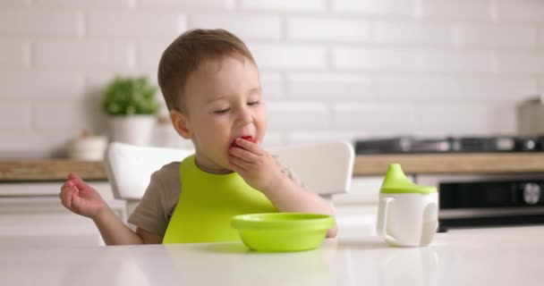 Roztomilý chlapeček sedí u stolu v kuchyni a rukou jí jahody ze zeleného talíře — Stock video