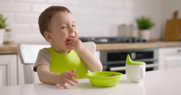 Roztomilý chlapeček sedí u stolu v kuchyni a rukou jí jahody ze zeleného talíře. Zpomalený pohyb. — Stock video