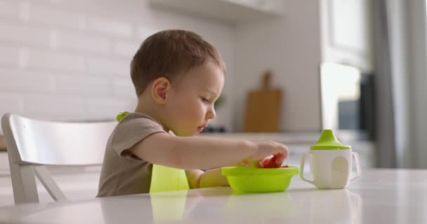 Roztomilý chlapeček sedí u stolu v kuchyni a rukou jí jahody ze zeleného talíře. Zpomalený pohyb. — Stock video