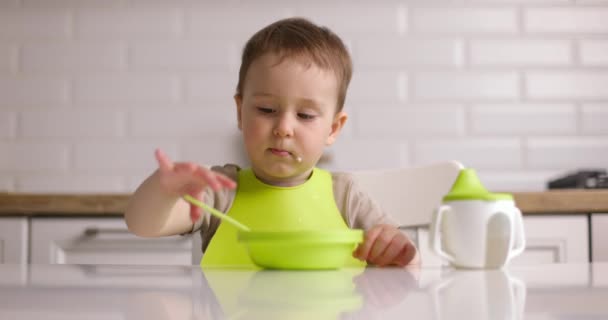 Nagyítás lövés a kis aranyos kisfiú ül az asztalnál, és eszik zabkását egy kanállal. A baba szívesen eszik otthon. Fogantatás Boldog gyermekkort — Stock videók