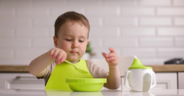 Egy aranyos kisfiú ül az asztalnál és zabkását eszik egy kanállal. A baba szívesen eszik otthon. Fogantatás Boldog gyermekkort — Stock videók
