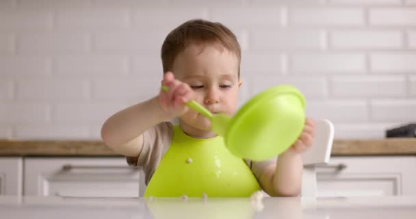 テーブルに座ってスプーンでオートミールを食べる小さなかわいい赤ちゃんの男の子。赤ちゃんは家で喜んで食べる。コンセプト｜Happy Yildary — ストック動画