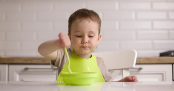 El niño desayuna en la cocina. El bebé feliz come gachas con cuchara. concepto de comer niño. — Vídeo de stock