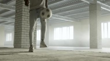 Genç siyahi adam içeride futbol topuyla antrenman yapıyor. Arkaplanda parlak güneş ışığı