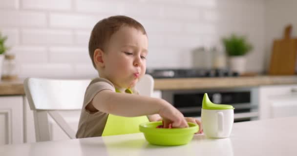 Το χαριτωμένο αγοράκι κάθεται στο τραπέζι στην κουζίνα και τρώει μούρα με το χέρι του. Η έννοια της ευτυχισμένης παιδικής ηλικίας και υγιεινής διατροφής. Αργή κίνηση. — Αρχείο Βίντεο