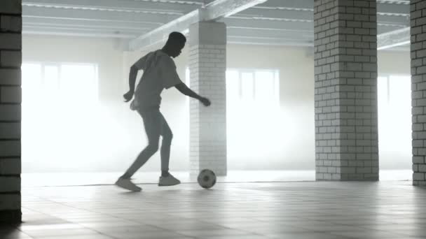 Молодой черный парень тренируется с футбольным мячом в помещении. Яркий солнечный свет на заднем плане — стоковое видео