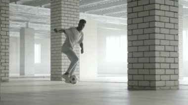 Genç siyahi adam içeride futbol topuyla antrenman yapıyor. Arkaplanda parlak güneş ışığı