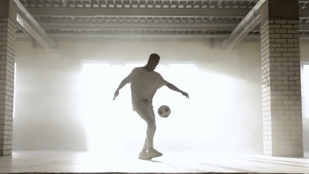 Joven negro entrena con una pelota de fútbol en el interior. Luz solar brillante en el fondo — Vídeos de Stock
