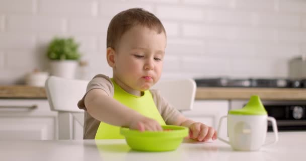 Το χαριτωμένο αγοράκι κάθεται σε ένα τραπέζι στην κουζίνα και τρώει φράουλες από το πράσινο πιάτο με τα χέρια του. Αργή κίνηση. — Αρχείο Βίντεο