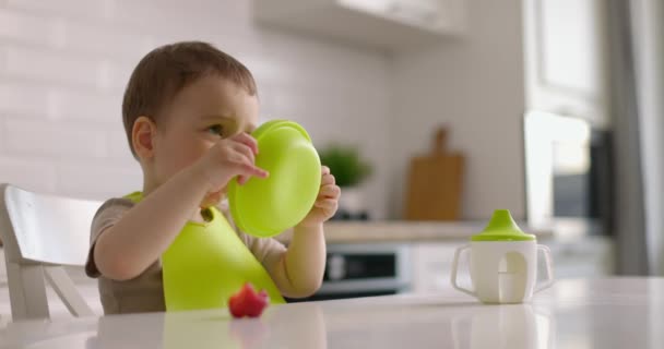 Roztomilý chlapeček sedí u stolu v kuchyni a rukou jí jahody ze zeleného talíře. Zpomalený pohyb. — Stock video