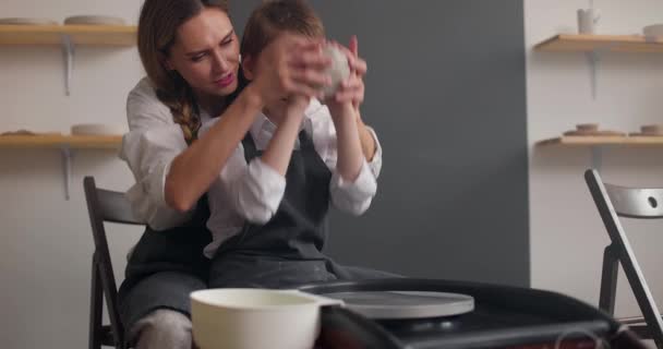 Bela mãe jovem e seu filho menino bonito fazendo peça de argila na roda de oleiro em estúdio. Movimento lento — Vídeo de Stock