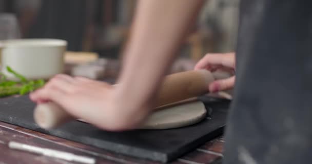 Manos femeninas halagando pieza de arcilla acostada sobre tela usando rodillo de madera en estudio de cerámica. Primer plano, cámara lenta — Vídeos de Stock